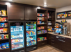 A hotel pantry with shelves stocked with snacks, drinks in a fridge, a coffee machine, and breakfast options.
