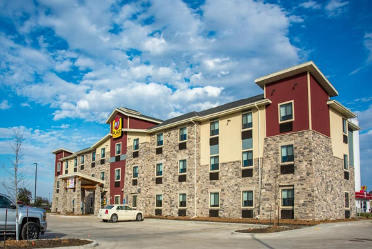 Four-story hotel with a mix of brick and stucco exterior, featuring a few parked cars in the lot and a clear blue sky above.