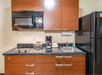 A kitchenette with brown cabinets, a small stove, sink, coffee machine, microwave, and a black refrigerator.