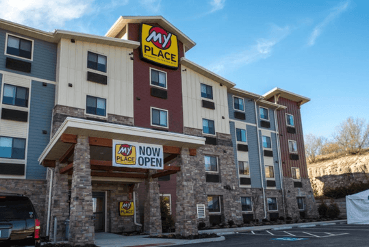 The front exterior of the My Place Hotel-Shakopee with blue, red, and white siding with stone accents and a "Now Open" sign up.