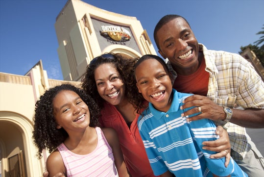 A smiling family posing together in front of a theme park entrance under a clear blue sky.