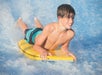 A boy wearing swim trunks rides a yellow bodyboard on water, surrounded by splashes, with a smile on his face.