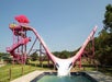 A bright pink water slide with a steep V-shape ends in a pool; two people are riding tubes down the slide on a sunny day.