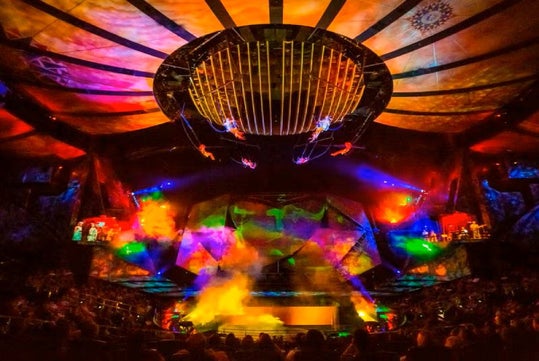 Colorful stage with dramatic lighting, suspended performers, and smoke effects in front of an audience during a live show.