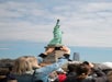 A crowd of people faces the Statue of Liberty; a person in the foreground raises a phone to take a photo. The sky is partly cloudy.