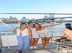 Three women pose and smile on a boat with arms raised, with a cityscape and a bridge over a river in the background.