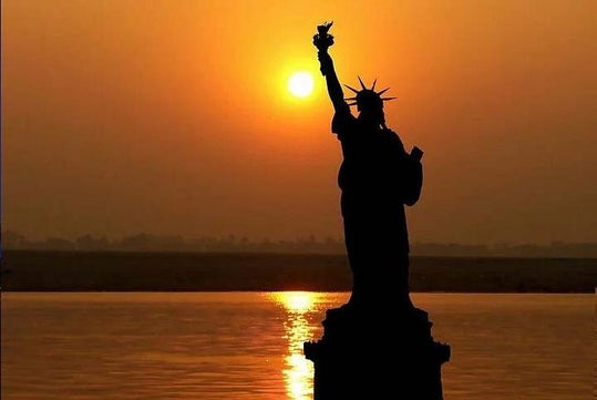 Silhouette of the Statue of Liberty against a sunset, with the sun reflecting on the water.