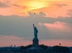 The Statue of Liberty is silhouetted against a sunset sky with clouds. An airplane and helicopter are visible in the distance.