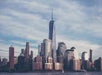 Skyline of a city with tall modern buildings, dominated by a prominent skyscraper with a spire against a partly cloudy sky.