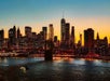 Silhouette of a city skyline with illuminated buildings against an orange and blue sunset sky, viewed across a river with a boat.