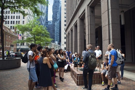 A group of people gathered on a city sidewalk, listening to a speaker. A tall building and trees are in the background.