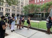 A group of people wearing masks stand in a line on a sidewalk beside a grassy area in front of buildings.