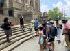A group of people wearing masks stand near the steps of a large stone building, listening to a guide.