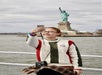 A person in a sweater takes a selfie on a boat with the Statue of Liberty visible in the background.