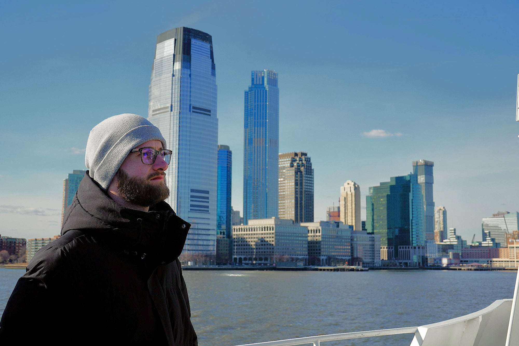 A man in a winter coat and beanie stands on a boat with modern city skyscrapers and a river in the background.