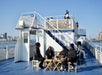 Several people sit and converse around tables on the deck of a ferry boat, with others on the upper level and a city skyline visible in the background.