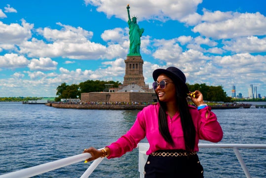 A woman in a pink shirt and black hat stands on a boat near the Statue of Liberty, with blue sky and clouds in the background.