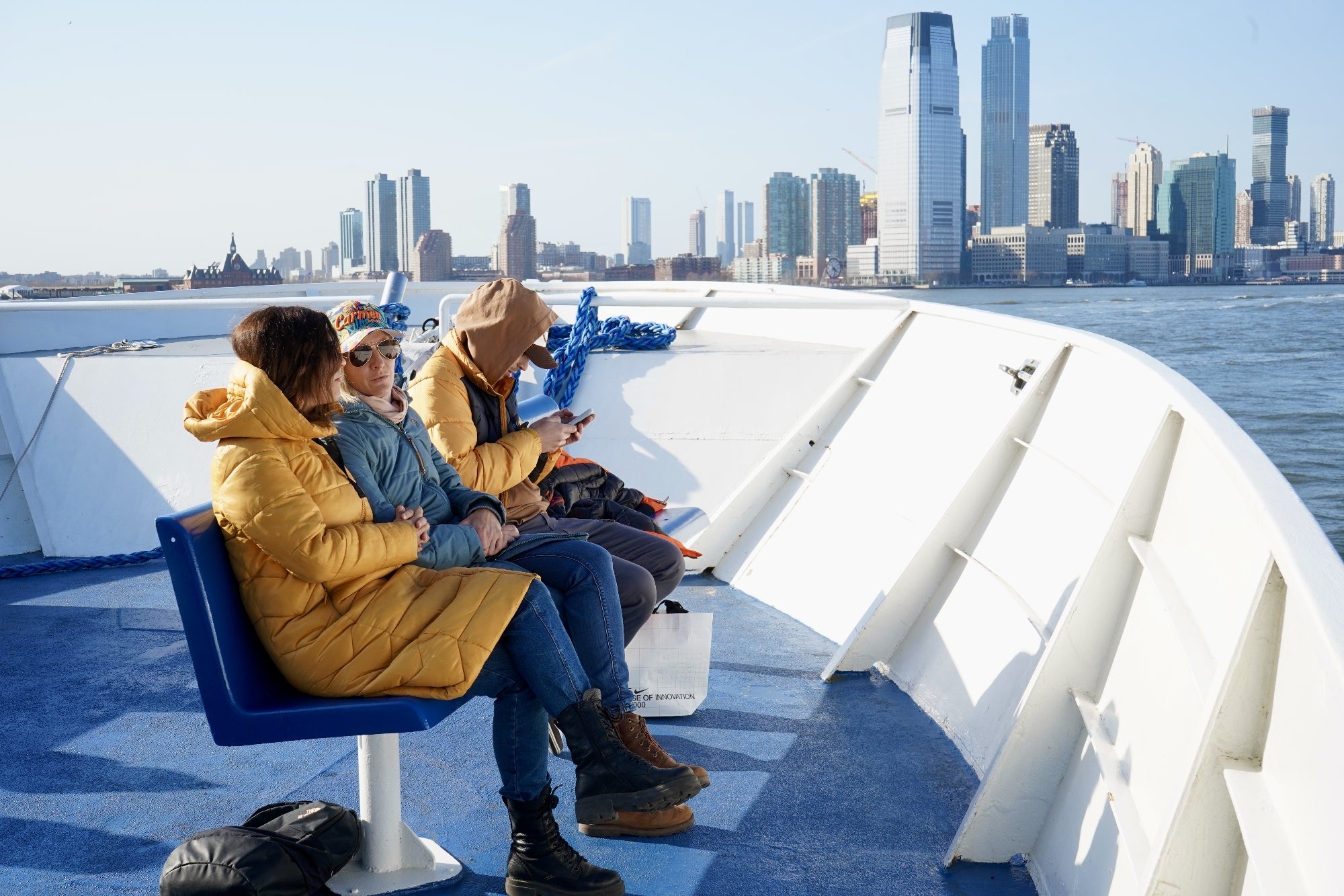 Three people sitting on a blue bench on a ferry, wearing jackets, with a city skyline and waterfront buildings in the background on a clear day.