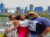 Three people pose and smile together outdoors with a city skyline and waterfront in the background.