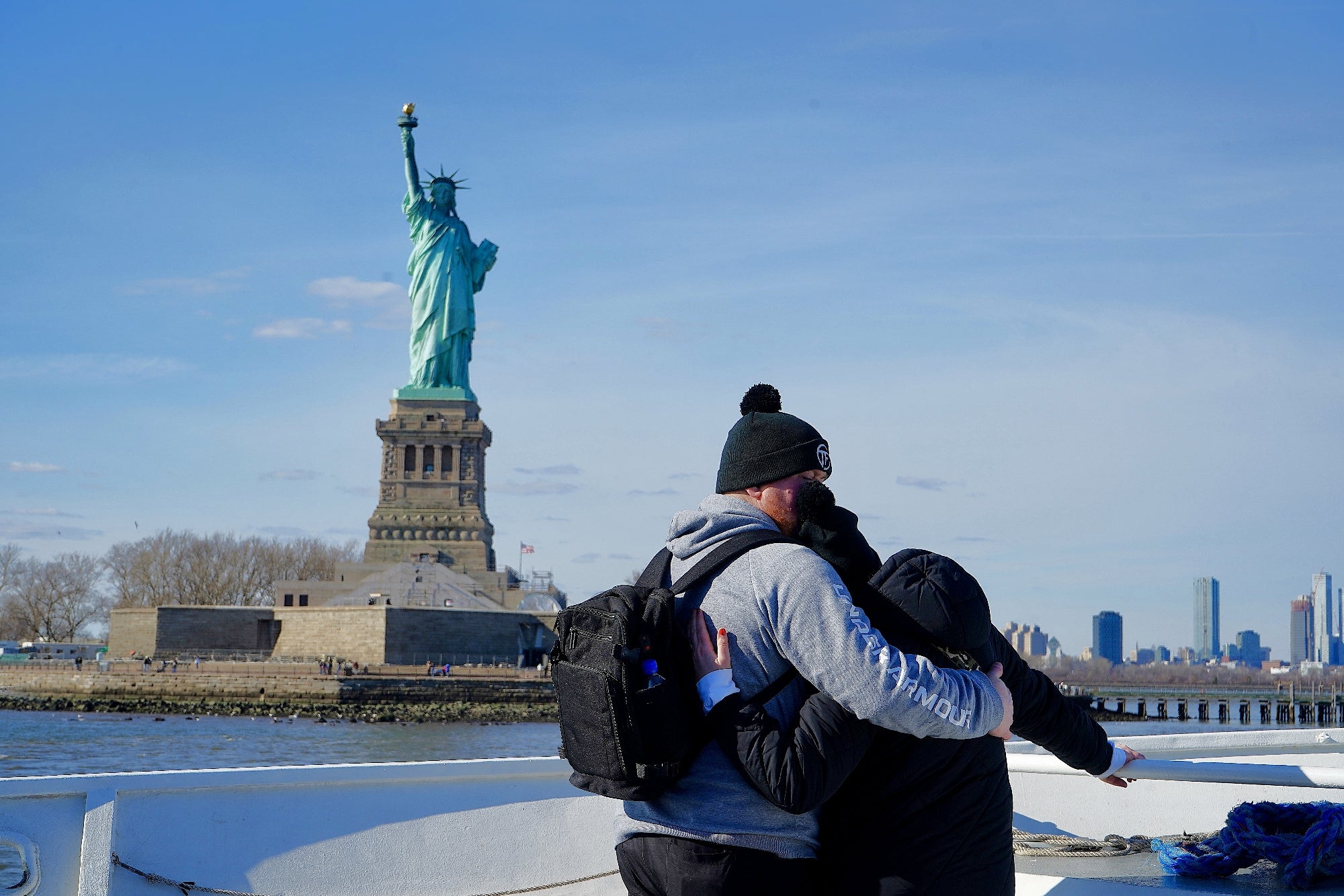 Two people stand on a boat, hugging and looking toward the Statue of Liberty with the New York City skyline visible in the background.