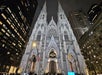 St. Patrick’s Cathedral in New York City at night, lit up, with American and Vatican flags at the entrance, surrounded by tall buildings.