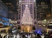 A large, brightly lit Christmas tree stands above the ice skating rink at Rockefeller Center in New York City, with people skating and city buildings in the background at night.