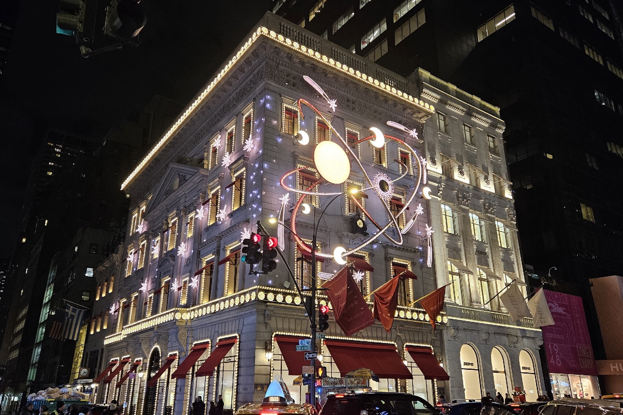 A brightly lit, decorated building at night, featuring red awnings, illuminated window displays, and large atomic-themed holiday lights on its facade. Traffic and street signs are visible in front.