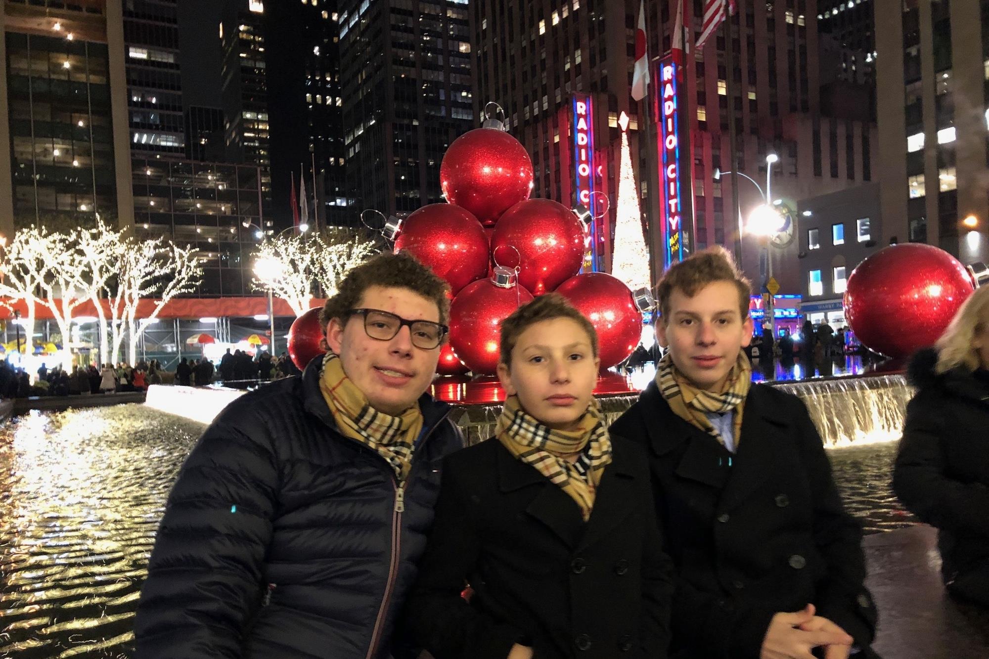 Three boys in matching scarves pose in front of a large Christmas ornament display and fountain near Radio City Music Hall in New York City at night.