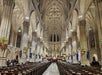 Interior view of a large cathedral with vaulted ceilings, rows of wooden pews, people seated, and festive wreaths hung on stone columns.