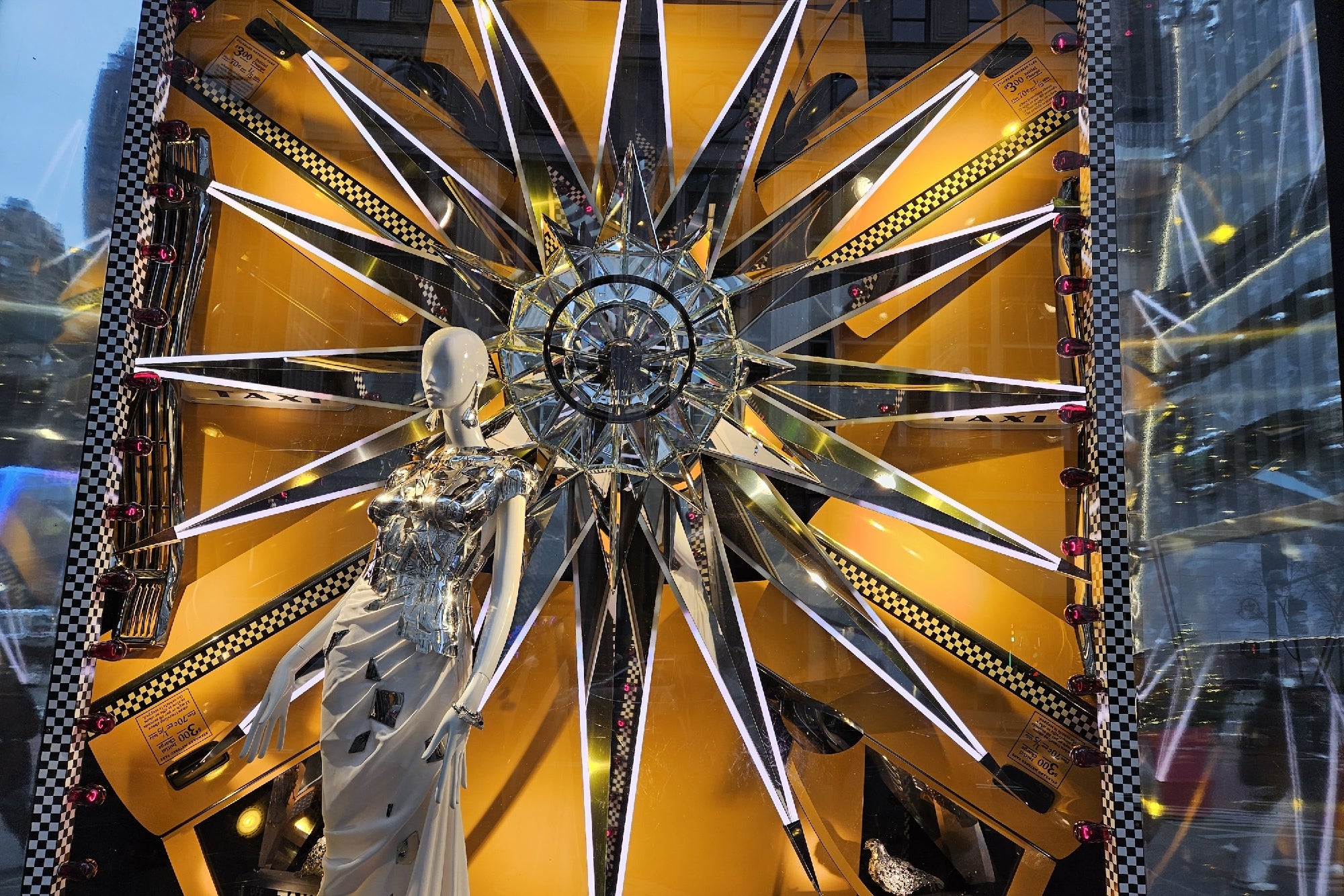A mannequin in a geometric silver dress stands before a large starburst display with mirrored spokes, set against a yellow background with small checkered details and toy cars.