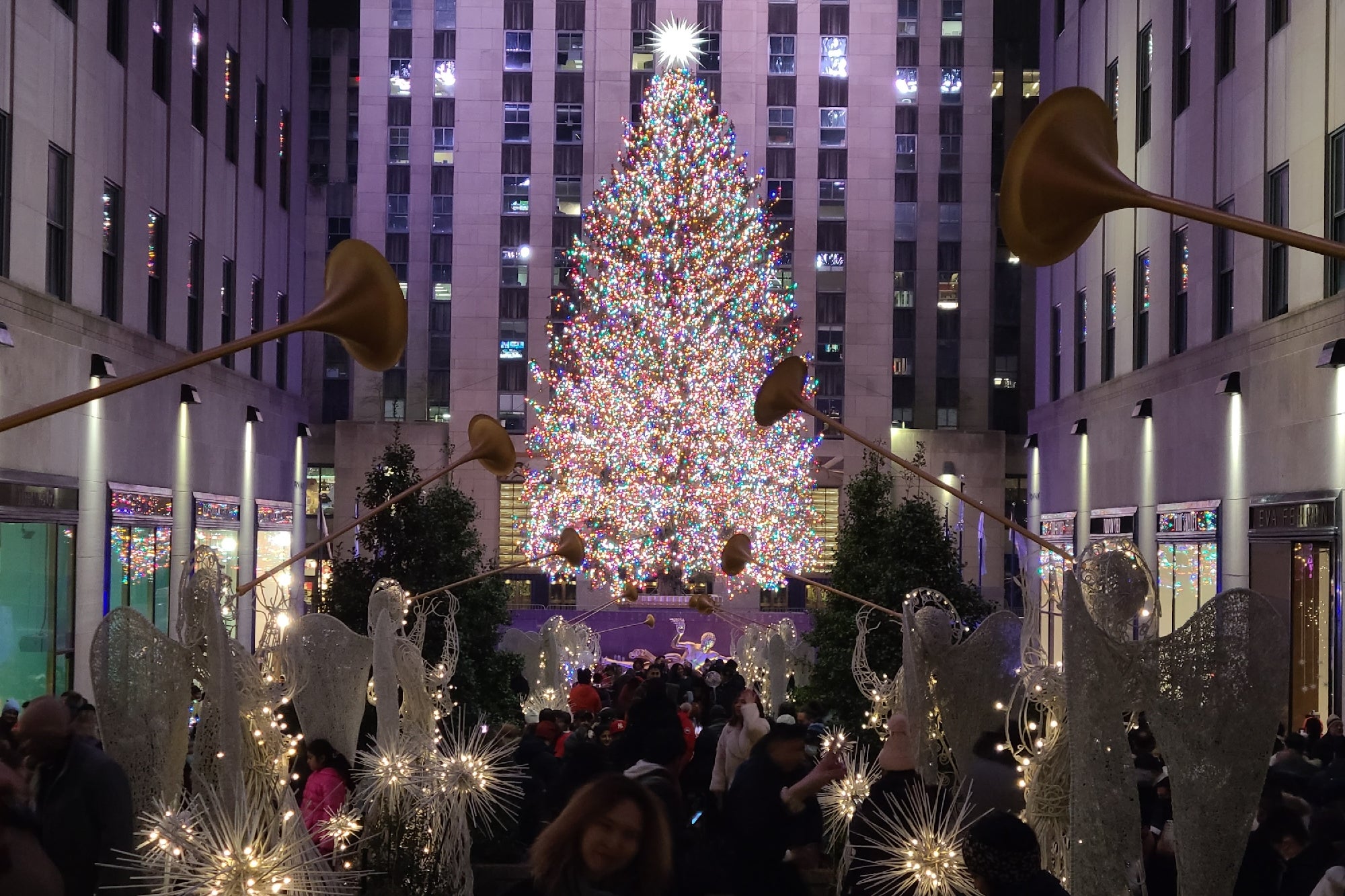 Crowd gathered at night in front of a large, brightly lit Christmas tree at Rockefeller Center, surrounded by angel decorations and festive lights.