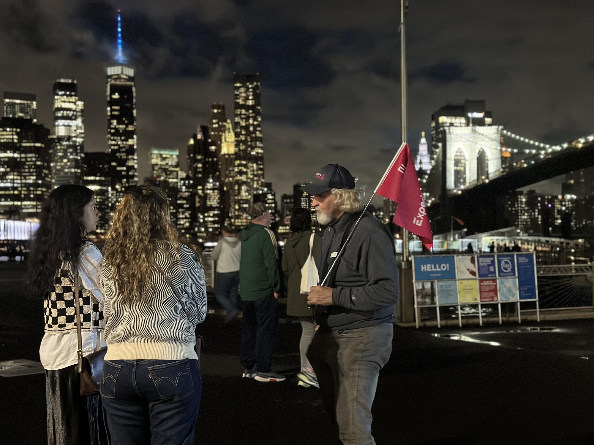 A man holding a red flag talks to two women at night with a city skyline and Brooklyn Bridge in the background.