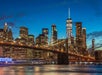 The Brooklyn Bridge spans across the East River at dusk, with the illuminated Manhattan skyline and One World Trade Center in the background.