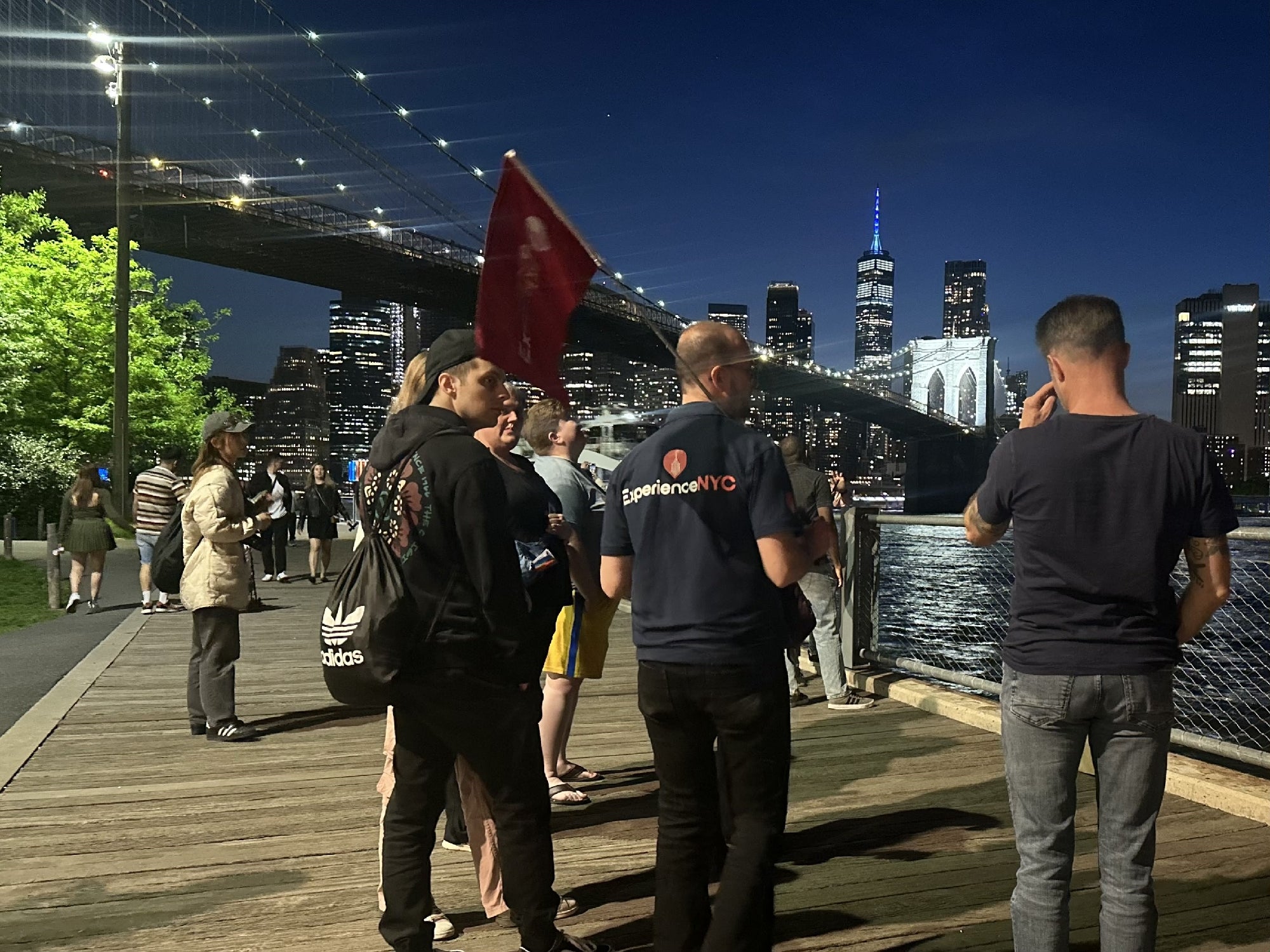 A group of people stands by the waterfront at night near the Brooklyn Bridge, with city buildings lit up in the background.