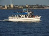A white double-deck boat named Karin Lynn carries passengers on calm water near a shoreline with buildings and palm trees in the background.