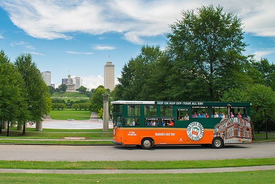 An orange and green tour trolley bus drives through a park with passengers, city buildings, and a clear blue sky in the background.