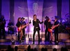 A male singer in a black jacket performs on stage with two female dancers, a live band, and purple lighting in the background.