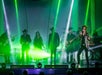 A band performs on stage with green laser lights and smoke effects, featuring multiple musicians including a vocalist at the front and a keyboard player in the background.