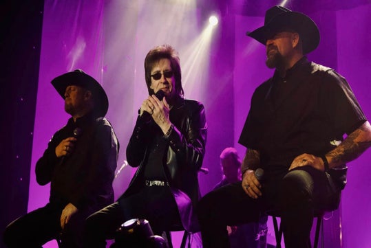 Three men wearing dark clothing and hats sit on stools, holding microphones and performing on a stage with purple lighting in the background.