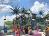 People enjoy a colorful outdoor water park with slides, water features, and fake palm trees under a partly cloudy sky.
