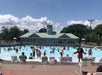 Outdoor swimming pool with many people in the water and several lifeguards present; lounge chairs line the poolside; a building is in the background under a partly cloudy sky.