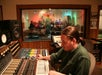 A man operates a large audio mixing console in a recording studio control room, with a group of people visible singing in the recording booth through a window.