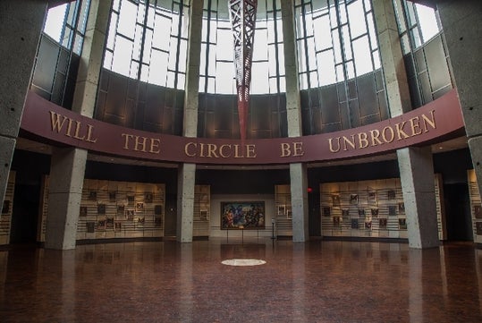Interior view of a rotunda with large windows and a banner reading "WILL THE CIRCLE BE UNBROKEN" above a display of photos and artifacts on the walls.