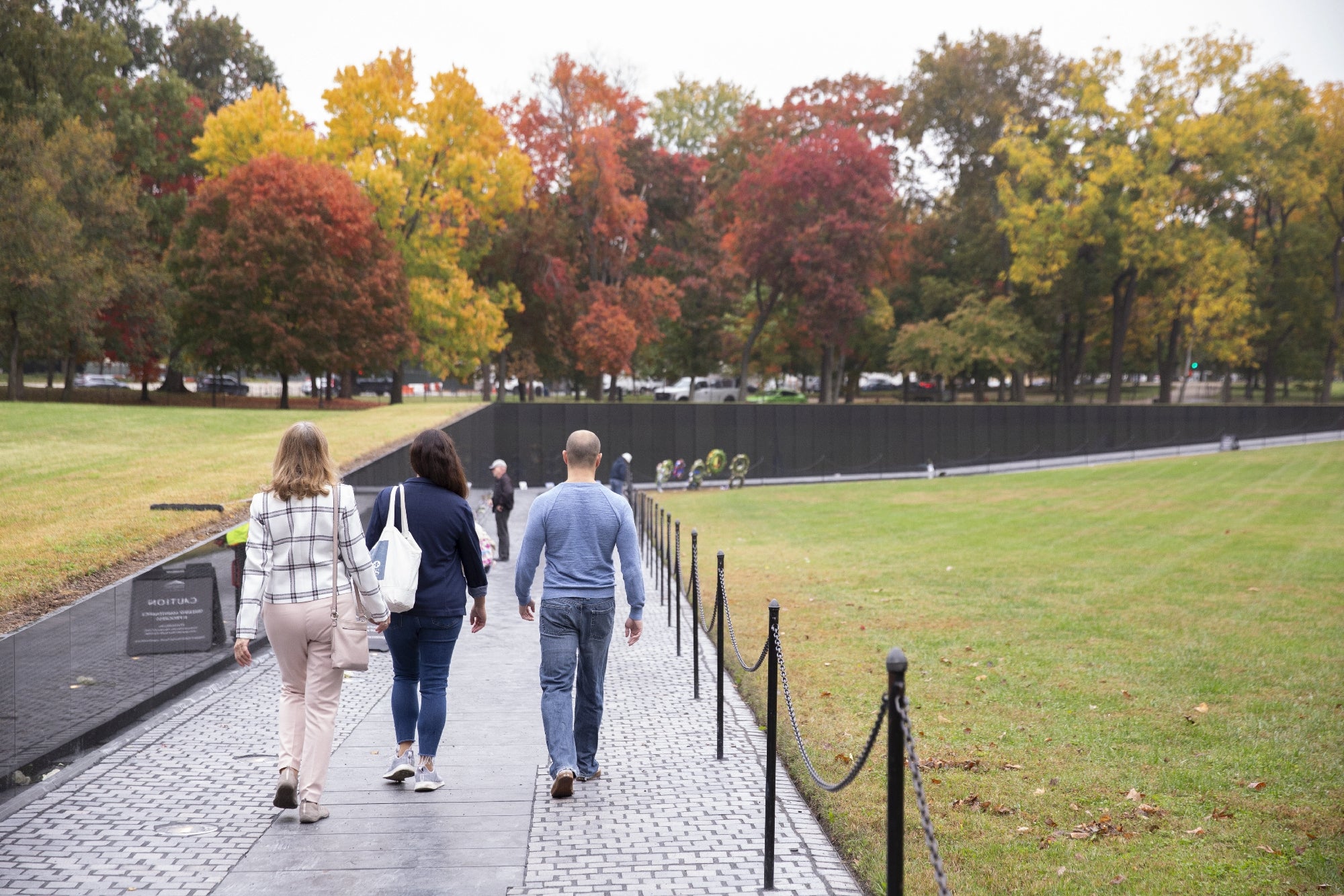 Three people walk along the path at the Vietnam Veterans Memorial in Washington, D.C., with autumn trees in the background.