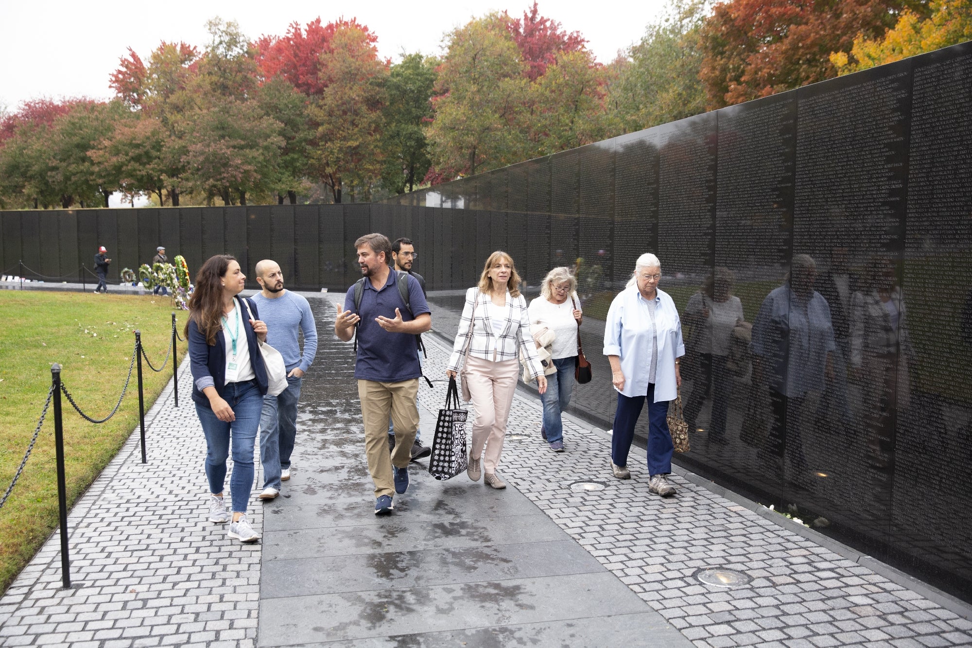 A group of people walk along the Vietnam Veterans Memorial wall, viewing engraved names, with autumn trees in the background.