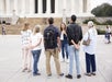A group of six adults stands in front of the Lincoln Memorial, listening to a woman speaking, with wide steps and columns in the background.