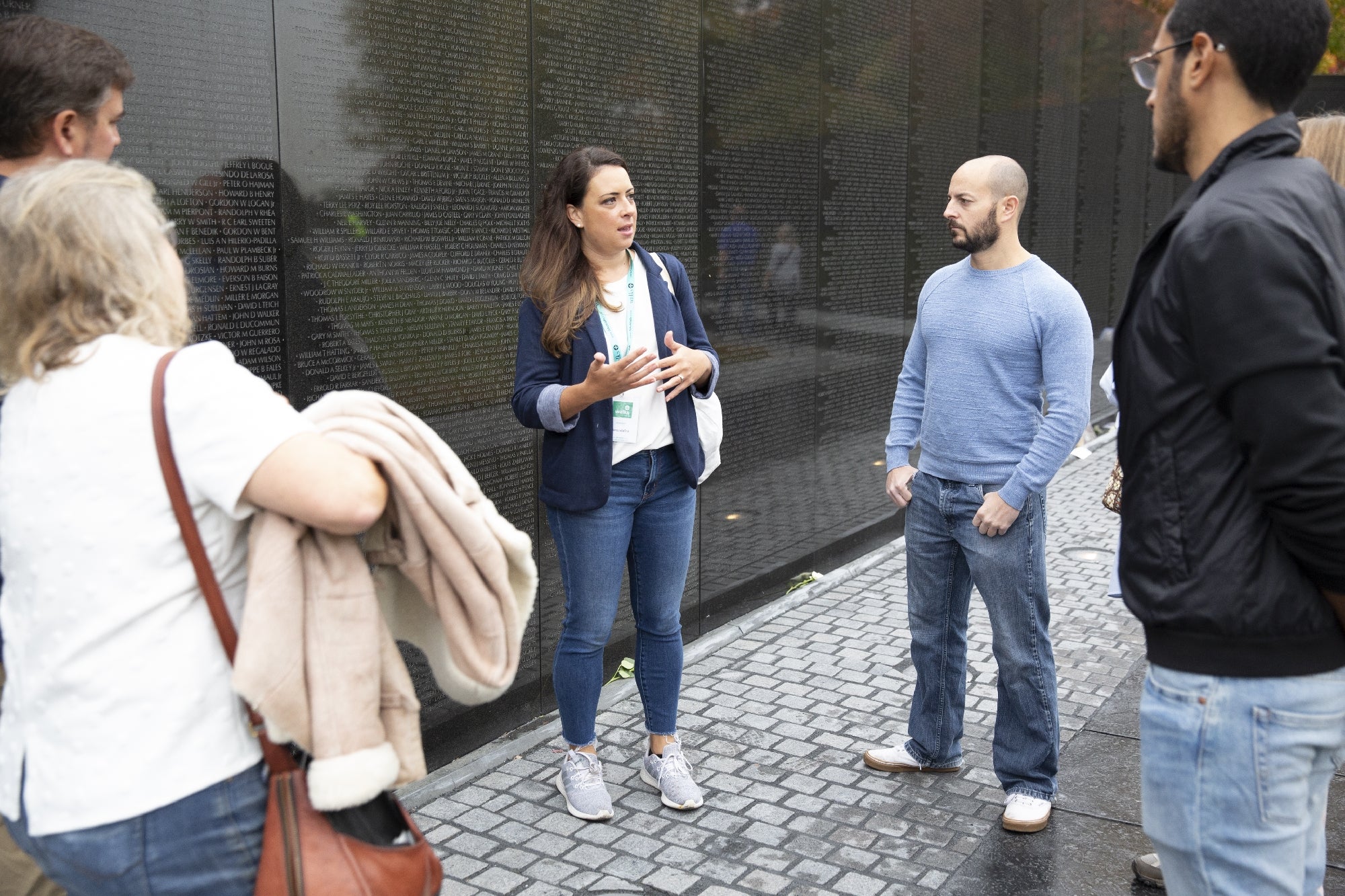 A group of people stand and talk in front of a black granite wall engraved with names, likely at a memorial site.