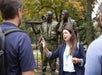 A woman speaks to two people outdoors in front of a bronze soldier statue, with trees in the background.