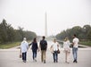 Six people walk towards the Washington Monument on a cloudy day, with trees and the reflecting pool visible on either side.