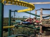 A person wearing safety gear rides a zipline near a tall wooden and metal structure, with trees and blue sky in the background.